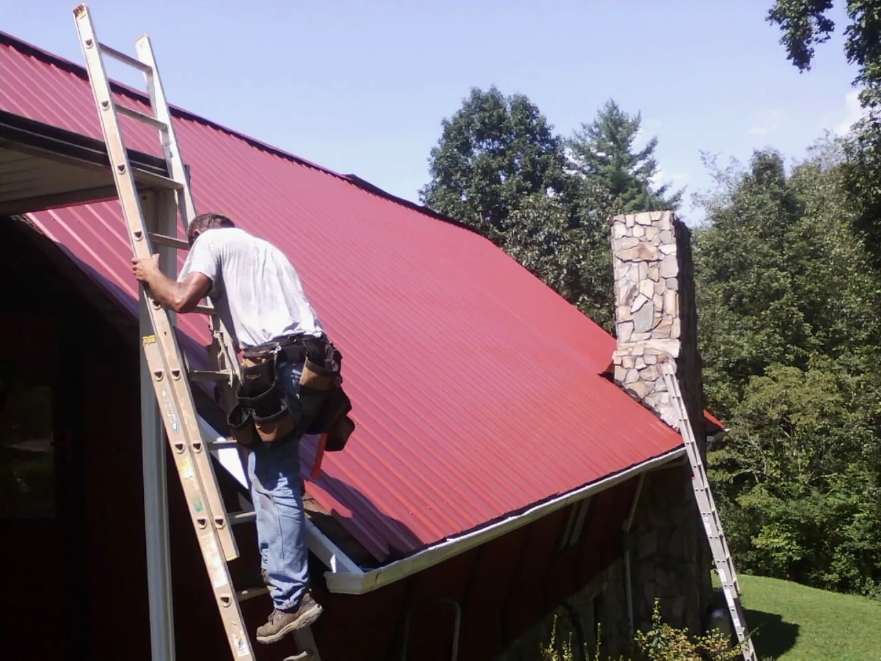 Roofer on a ladder beside a red metal roof and stone chimney — Soffit & Fascia Repair by Roofing Brothers in Claremont